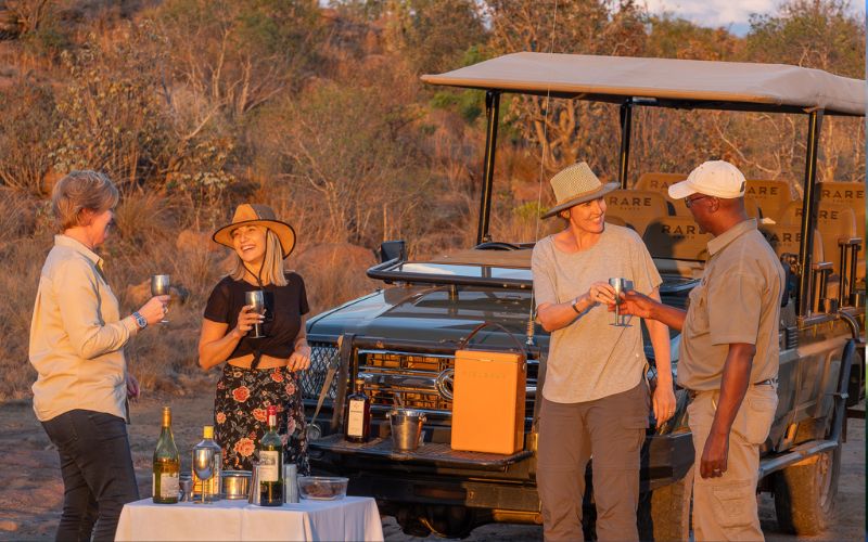 Sian Loehrer and Vikki Jackson of Marula Hill Travel enjoying a safari sundowner beside a game-viewing vehicle in South Africa’s bushveld.
