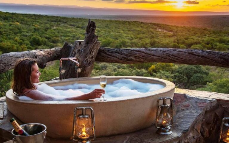 A guest enjoys a bubble bath with a glass of champagne overlooking the African wilderness at sunset, embracing ultimate safari luxury.